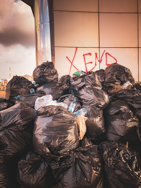 A worker wearing a yellow and red uniform is operating a large red rubbish collection truck parked on the side of a street. The truck's rear opening is exposed, showing mechanical components and a loa