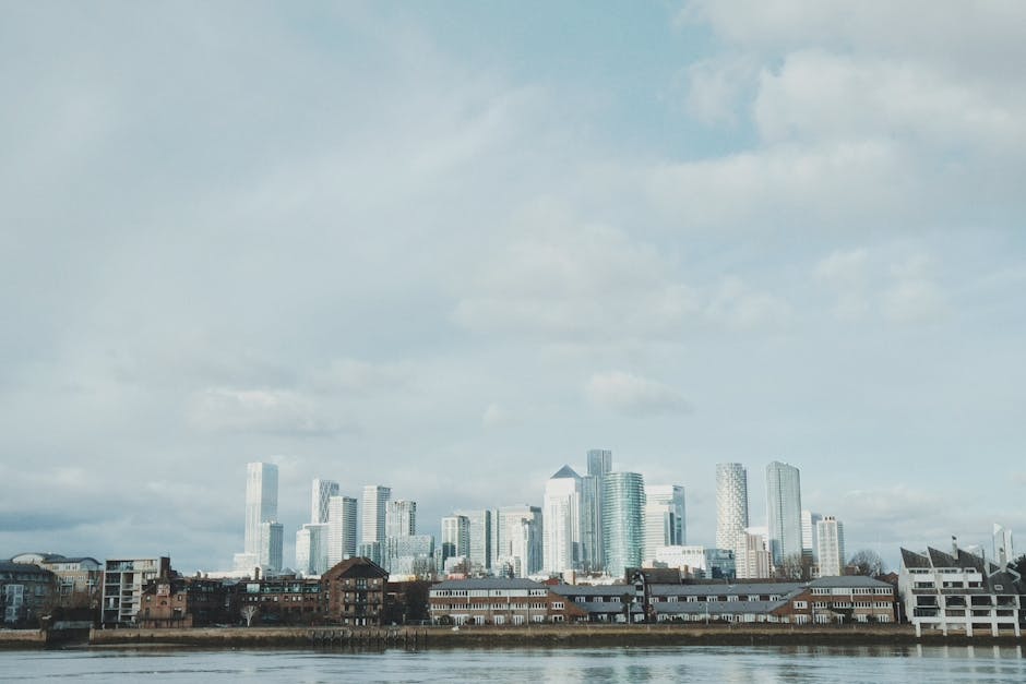 A panoramic view of a modern cityscape during dusk, featuring a variety of high-rise office buildings with reflective glass facades and unique architectural designs. In the foreground, there is a tall