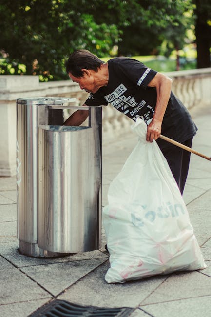A large collection of overflowing rubbish bins and discarded waste bags, including cardboard boxes, paper, plastic packaging, and black garbage bags, situated on a paved sidewalk in front of a commerc