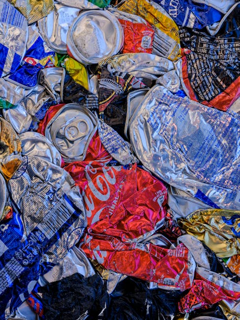 A close-up view of a pile of crushed aluminium beverage cans, primarily in silver, with some featuring printed labels in red, blue, yellow, and black. The cans are crumpled and stacked irregularly, wi